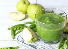 green smoothie in a clear glass sits on a rustic white counter with sliced and whole apples and open pea pods sitting next to it